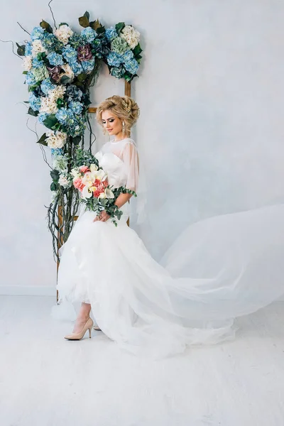 Gyönyörű menyasszony, menyasszonyi ruha és cipő a csokor a kezében, a háttérben a falra virággal, vonat kialakítása — Stock Fotó