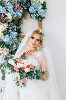 Duvak içinde bir gelin portresi duruyor onun elinde çiçeklerle beyaz bir duvara