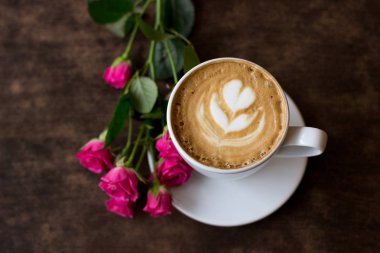 Beyaz bir fincan sıcak, hoş kokulu kahve köpüklü bir kalp şeklinde bir gül dalı yalan yanında koyu ahşap arka plan üzerinde duruyor. metin için yer