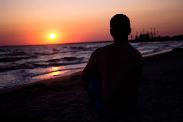 Silhouette eines Mannes, der auf dem Sand am Meer sitzt, in den Strahlen der untergehenden Sonne am Meer. unter den Text legen, sich am Meer entspannen — Stockfoto