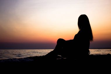 gün batımında deniz kenarında yaz aylarında genç kız gün batımını izlemek kum üzerine oturur. gün batımında siluet