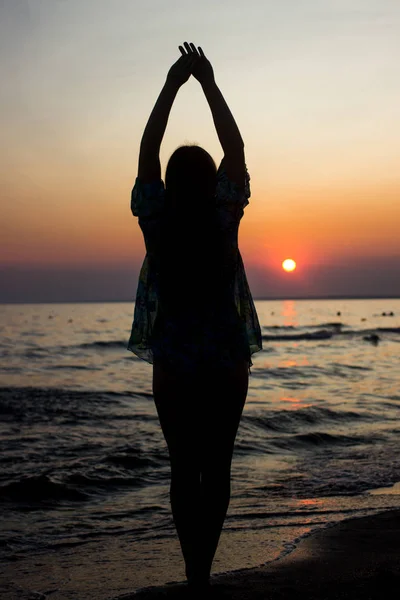 silhouette of a young, slender girl in the background of the setting sun on the beach, raised hands up, elongated silhouette