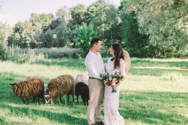 Gelinlik giymiş genç ve güzel bir çift yeşil bir çayırda duruyor, arka planda sığırlar var.