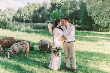 Gelinlik giymiş genç ve güzel bir çift yeşil bir çayırda duruyor, arka planda sığırlar var.