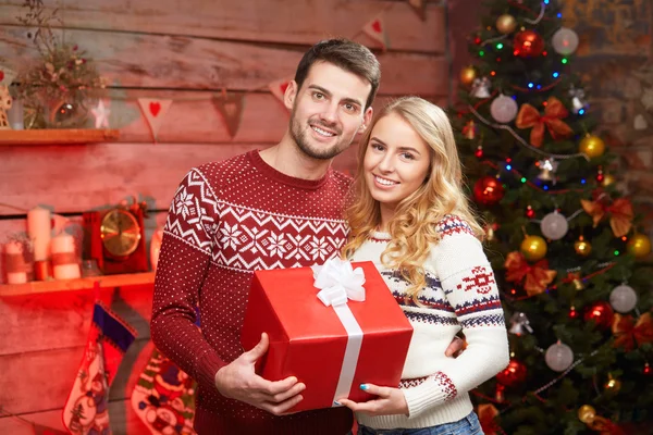 Jovem casal em camisolas abraçando e segurando caixa de presente vermelho — Fotografia de Stock