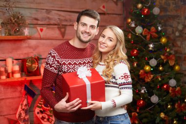 Happy Couple in winter sweaters smiling and holding big red gift box clipart