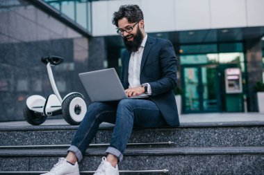 Gülümseyen işadamı elektrikli scooterla mermer merdivenlerde dizüstü bilgisayarda yazı yazıyor.