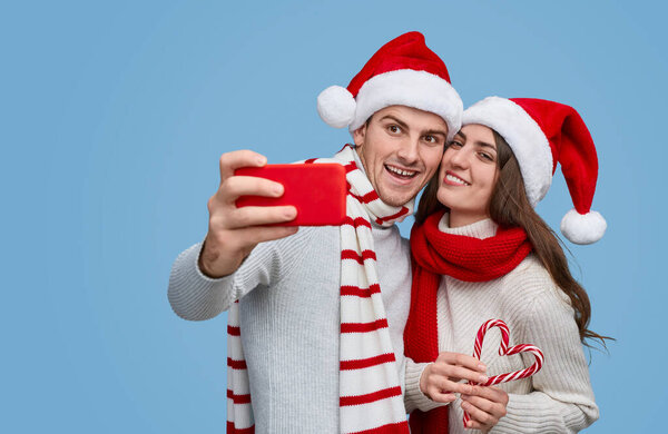 Loving couple in Santa hats taking selfie while carrying candy cane