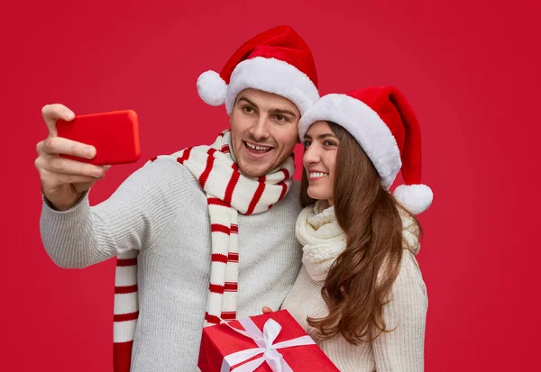 Casal feliz em chapéus de Papai Noel levando selfie enquanto carrega caixa de presente — Fotografia de Stock