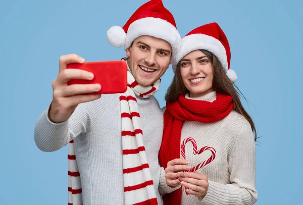 Casal alegre em chapéus de Papai Noel tirar selfie enquanto carrega cana de doces — Fotografia de Stock