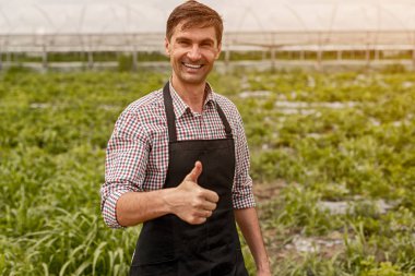 Siyah önlüklü gülümseyen bitki yetiştiricisi serada başparmağını kameraya gösteriyor.