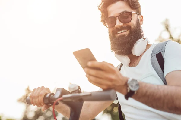 Man tersenyum sambil melihat-lihat smartphone pada skuter di jalan — Stok Foto