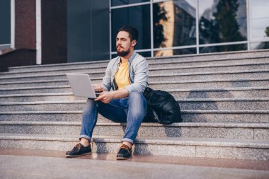 Sakallı öğrenci basamaklarda dizüstü bilgisayar kullanıyor.