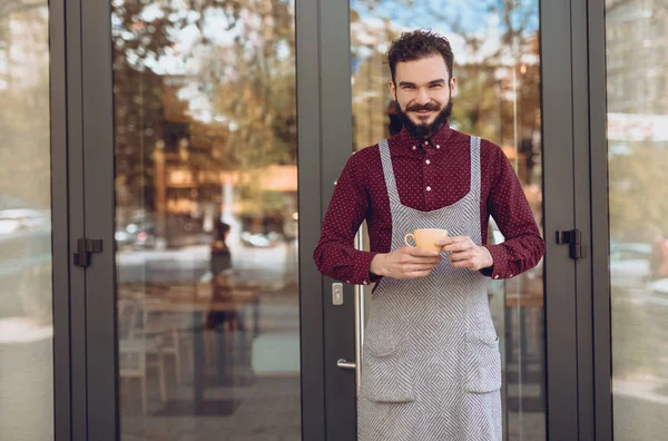 Amistoso camarero con taza de pie cerca de la entrada de la cafetería — Foto de Stock