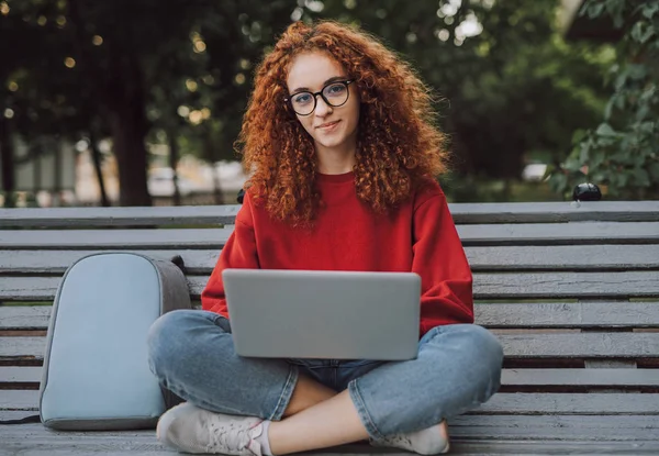 Positive young woman with laptop sitting on bench in city Stock Photo