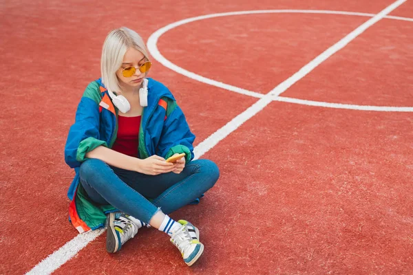 Pensive teen girl in yellow sunglasses using smartphone while resting on playground — Zdjęcie stockowe