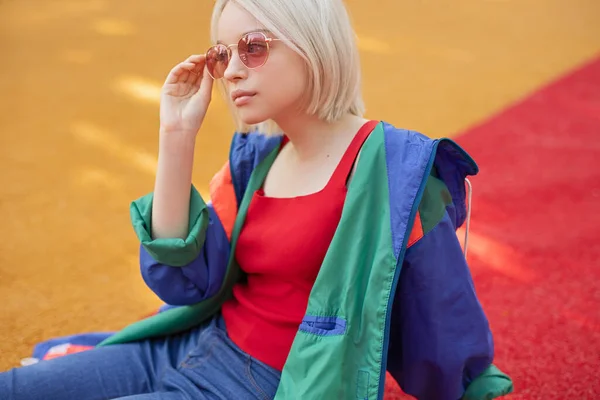 Menina adolescente confiante na moda olhando para longe e ponderando enquanto sentado no chão e tocando óculos rosa — Fotografia de Stock