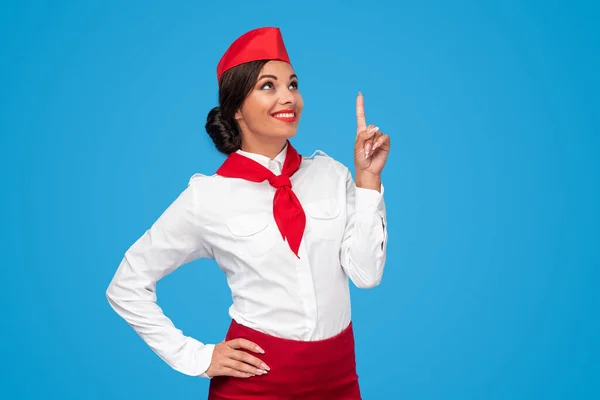 Прекрасная молодая стюардесса в красно-белой форме, указывающая вверх и улыбающаяся — стоковое фото