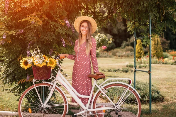 Young pleased woman in straw hat and glasses with bike in summer garden — Stok fotoğraf