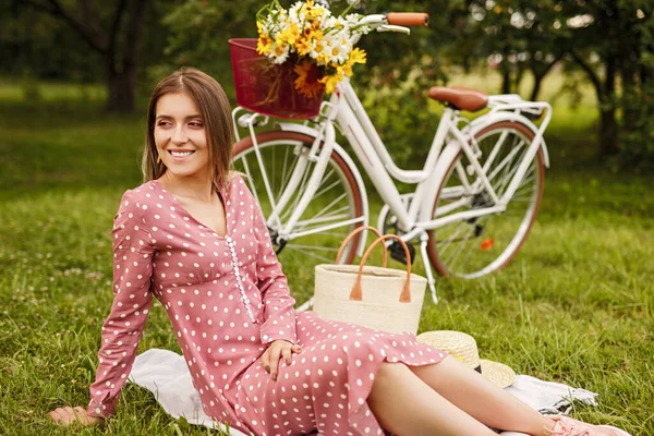 Cheerful adult woman sitting on ground beside bike while relaxing in summer garden — Zdjęcie stockowe