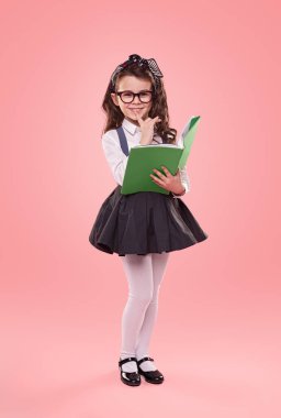 Gözlüklü, uzun boylu, zeki bir çocuk. Parmağını dudaklarının yakınında tutuyor ve pembe arka planda ders kitabı okurken gülümsüyor.