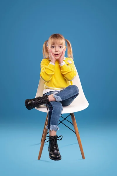 Surprised small girl in bright yellow sweater and jeans and black boots looking at camera and touching cheeks, while sitting alone on white chair against blue background in studio
