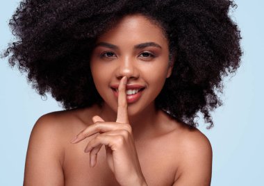 Kıvırcık saçlı güzel Afro-Amerikalı kadın kameraya bakıyor ve parmağını gülümseyen dudakların yanında tutuyor. Mavi arka plandaki güzelliğin sırları hakkında sessiz kalmak istiyor.