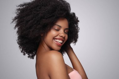 Kapalı gözlerle gülümseyen, temiz kıvırcık saçlara dokunan ve gri arka planda spa hizmetini temsil eden iyimser Afro-Amerikan bayanın yan görüntüsü