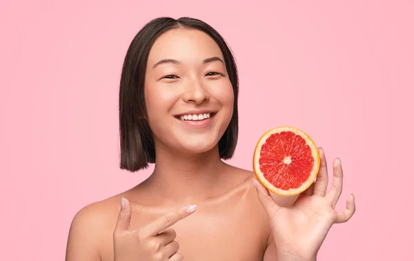 ピンクの背景に熟した赤いグレープフルーツを示す裸の肩を持つ幸せな健康的な若いアジアの女性 — ストック写真