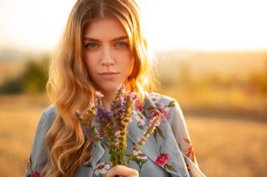 Sakin, genç, kızıl saçlı, şık elbiseli, elinde bir demet lavanta çiçeği tutan ve yaz tarlasında yanan kameraya bakan bir kadın.