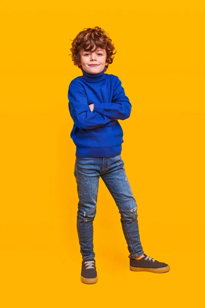 穿着蓝色毛衣和牛仔裤的积极自信的学童站在黄色的背景下看着相机 — 图库照片