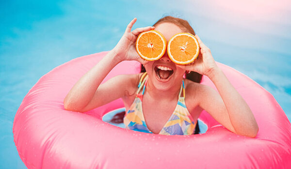 Cheerful active little girl in swimsuit covering eyes with sliced orange fruit while having fun in pool with pink inflatable ring