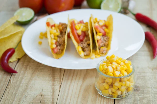 Yellow sweet corn in a small glass bowl and mexican tacos with chicken meat, chili pepper, tomato and corn as background — Stock Photo, Image