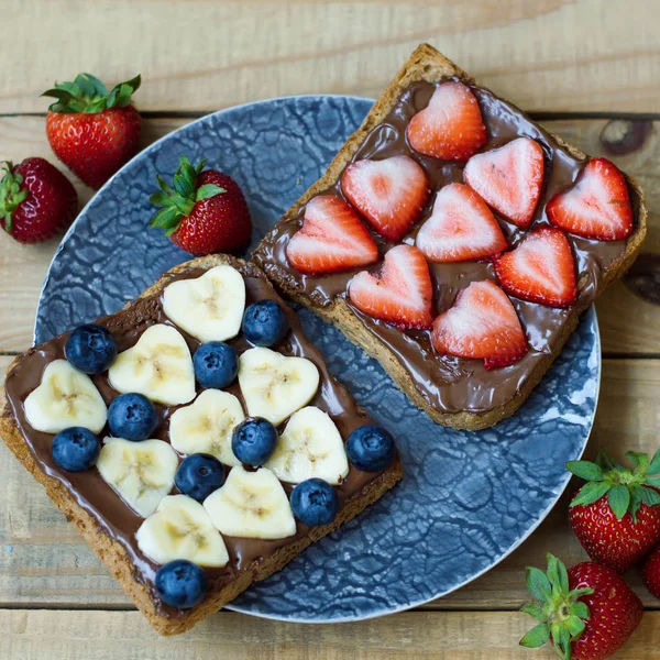 Square photo of two sweet toasts with heart shaped banana and strawberry, blueberry, hazelnut spread and whole grain bread