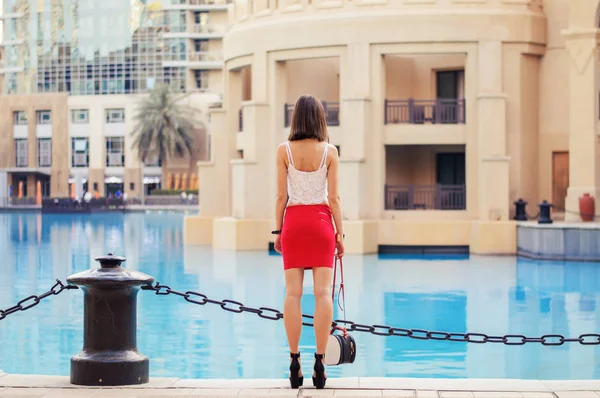Mujer europea flaca y alta en top blanco y falda roja corta con bolso pequeño y zapatos de tacón alto de pie cerca del paseo marítimo en el centro de Dubai — Foto de Stock