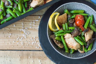 Üstten Görünüm sağlıklı sıcak fırında tavuk göğsü, yeşil fasulye, domates, salata, dilimlenmiş mantar, küçük bir hizmet limon ve susam tohumu kase ve kayrak tahta ahşap masa üzerinde yuvarlak