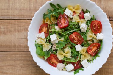 Kırmızı kiraz domates, beyaz peynir, farfalle, roka, Bebek ıspanak, ahşap masa üzerinde beyaz salata kaseye tavuk eti ile ev yapımı makarna salatası. Sağlıklı beslenme