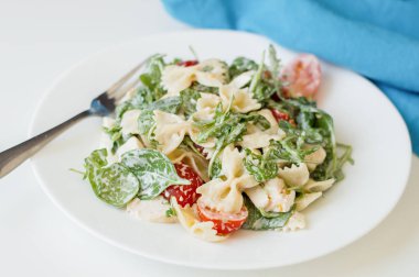 Makarna salatası rucola bırak, Bebek ıspanak, farfalle, dilimlenmiş domates ve tavuk eti ile bölümünü beyaz sos ile giyinmiş ve çatal ve mavi peçete ile beyaz tabakta servis