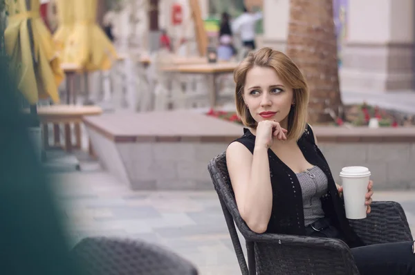 Horizontal photo of young european female sitting at the open air terrace with a paper cup of coffee. Lifestyle concept. Coffee break