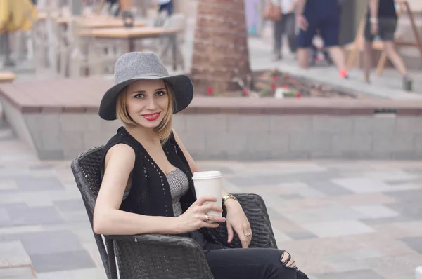 Porträtt av ung blond kvinna med bob skära frisyr, grå hatt, röda läppar och trendiga jeans sitter utanför och dricker kaffe. Leende och positiv kvinna med cappuccino — Stockfoto