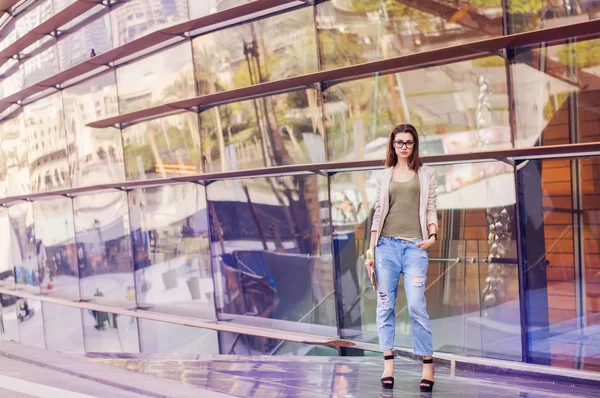 Young woman with bob hair cut, eyeglasses, in beige jacket and boyfriend jeans standing near glass building. Street fashion. Smart casual
