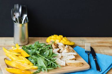 Pişmiş tavuk göğsü, mango dilimleri, roka, Bebek ıspanak ve badem dilimleri ahşap kesme tahtası mavi peçete, çatal ve bıçak ile servis. Dengeli yemek