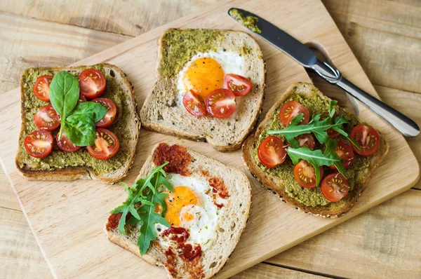 Kahvaltı tost ahşap kesme tahtası üzerinde. Kiraz domates, yumurta, ıspanak, roka, pesto sos, domates sosu ve susam ile ekmek ile sağlıklı sandviç. Dengeli kahvaltı