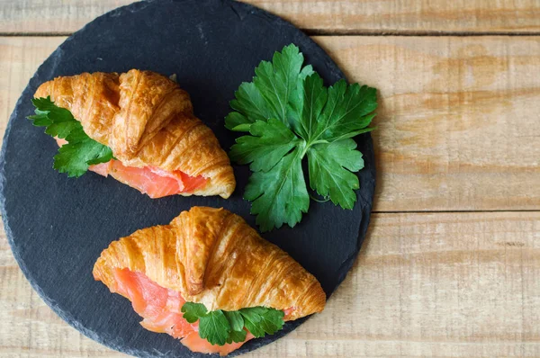 Croissant sandwiches with smoked salmon and parsley on black slate board on wooden table. Sandwiches with red fish and herbs. Breakfast meal — Stock Photo, Image