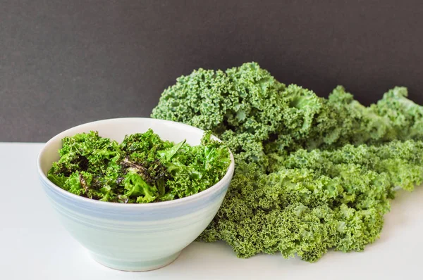 Bowl with crispy kale chips and fresh kale bunch on white table. Kale salad