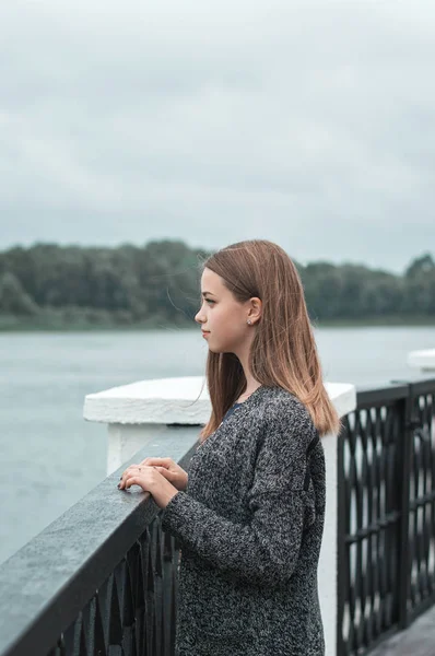 Vertikální portrét ženské teenagera v pletený kabát, stojící u mostu poblíž břehu řeky — Stock fotografie
