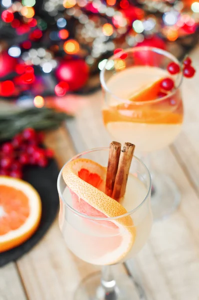 Cocktails with grapefruit and orange slices, cinnamon sticks and red currant served on wooden table with christmas decoration and lights as background. Party concept. Party drinks — Stock Photo, Image