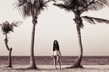 Uzun boylu egzotik palmiye ağaçları arasında kumlu Plajı'nda genç kadın ayakta. Siyah ve beyaz fotoğraf beyaz ıslak gömlek içinde kadın