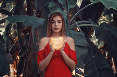 Büyük muz yaprakları duran ve parlak ve ışıltılı kalp tutan Kırmızı elbiseli kadın modeli portresi. Aşk sembolü