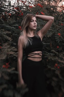 Vertical photo of a female blond model in black trendy jumpsuit standin near green bush with red flowers clipart